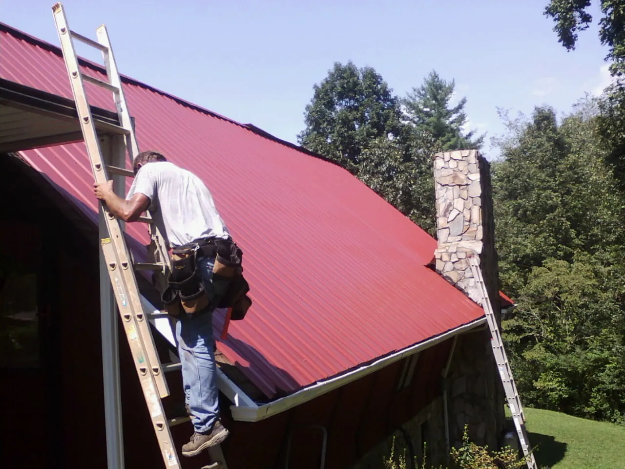 Roofer on a ladder beside a red metal roof and stone chimney — Soffit & Fascia Repair by Roofing Brothers in Smithfield